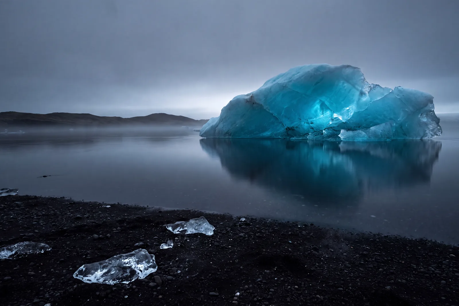 A blue-lit iceberg on a still lagoon beside a black volcanic beach.